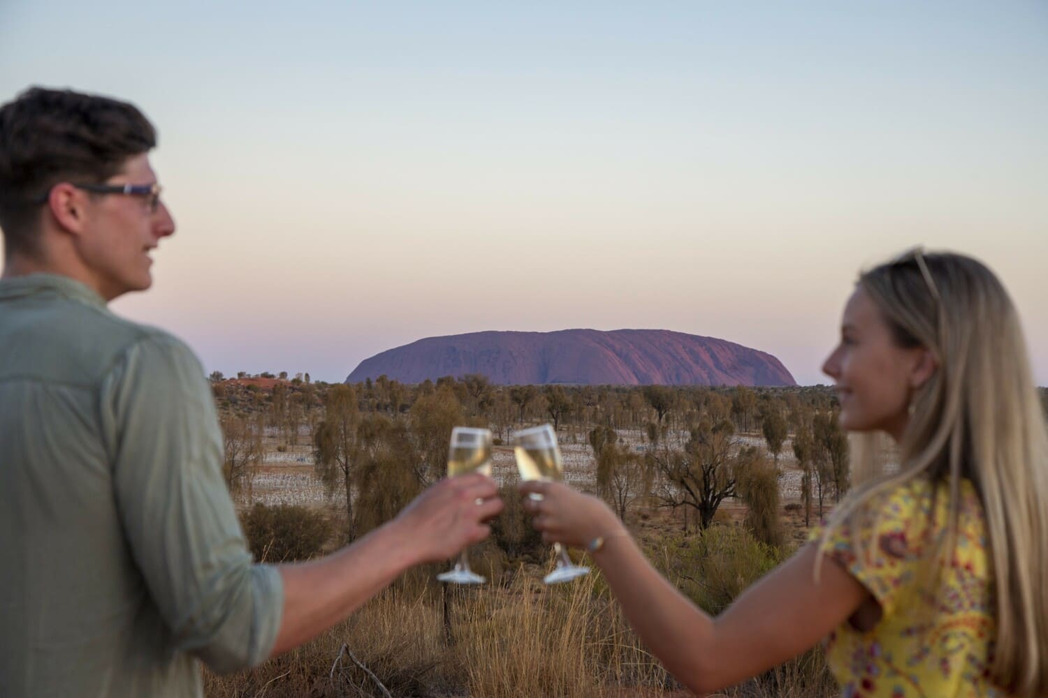 A man and a woman toasting with wine glasses in front of Uluru