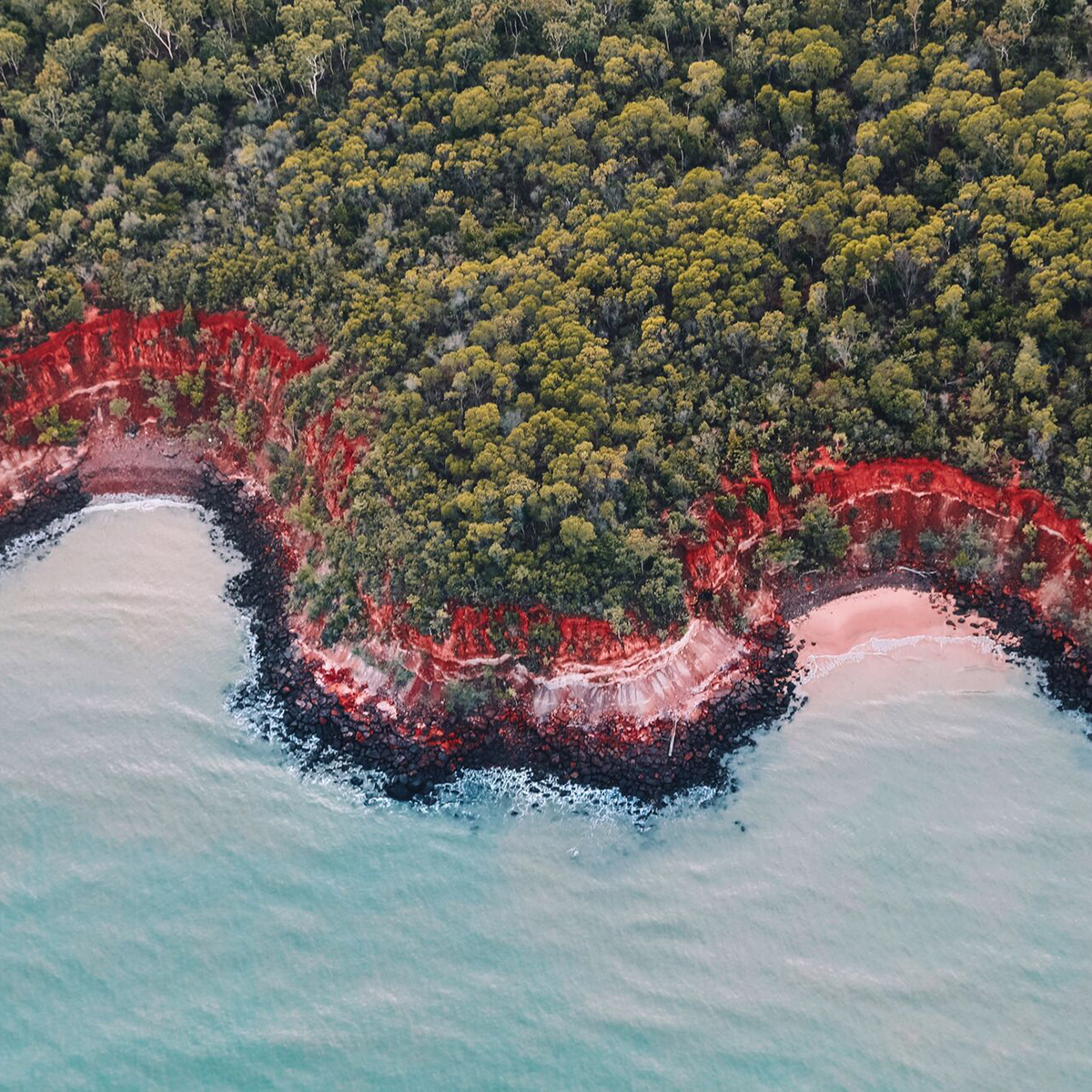 An aerial view of turquoise water, ochre red cliffs and verdant green trees on Tiwi Islands