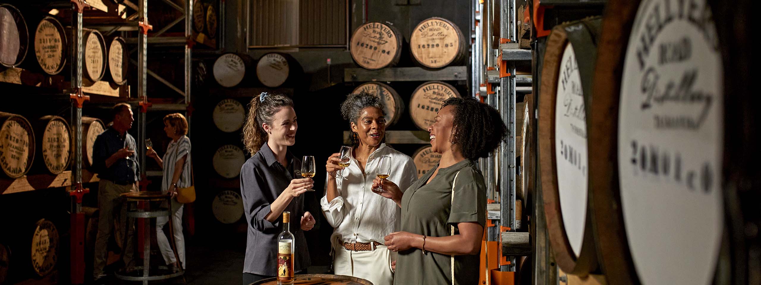 A group of people with glasses in their hands in a distillery with barrels in the background