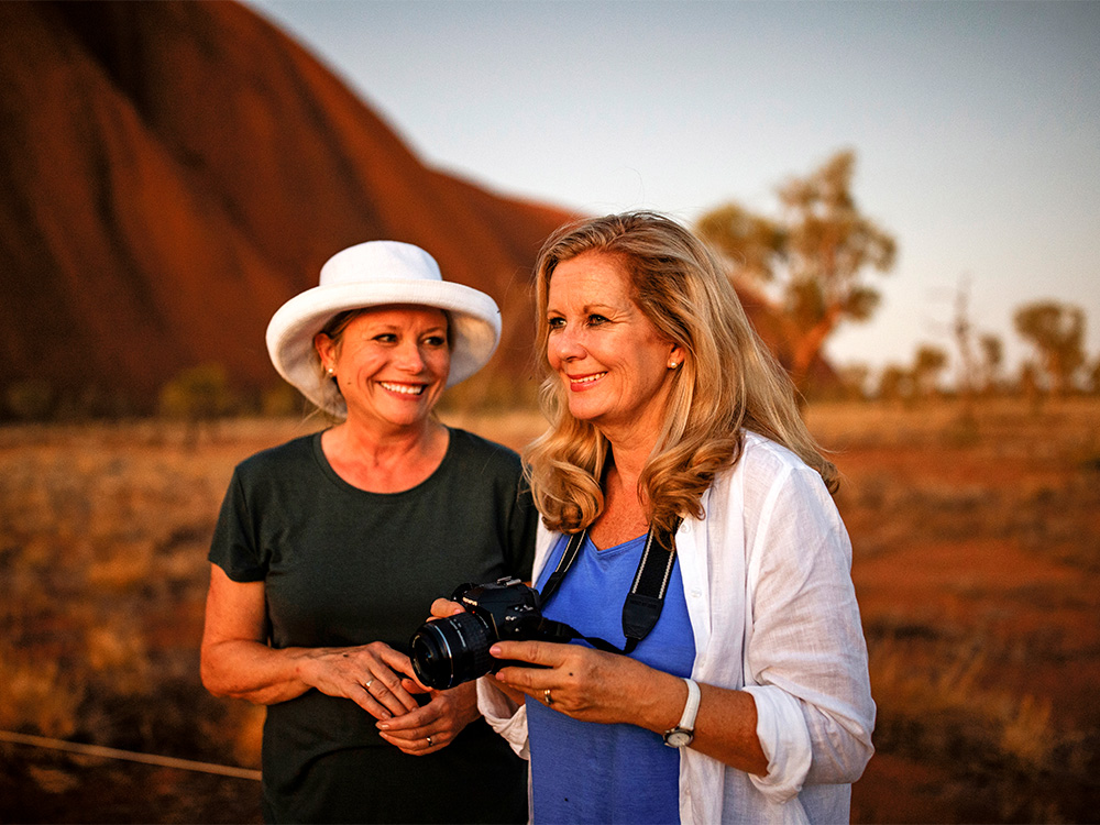 Woman with hat and woman with camera smile against red mountains
