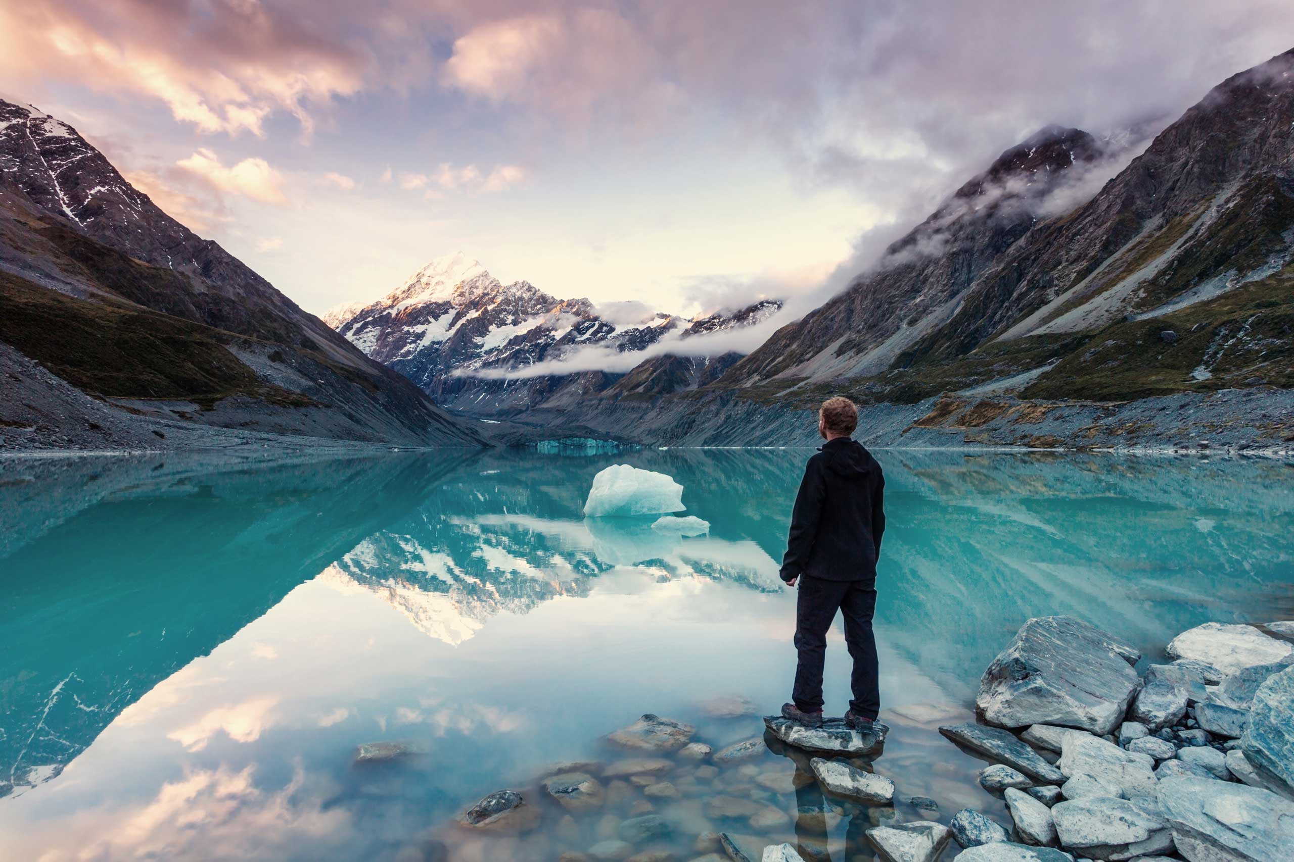 A man standing on a rock next to a body of water and view of the mountains