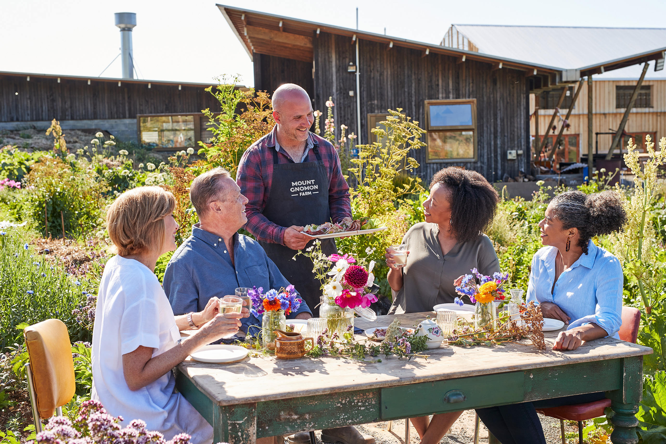  A group of people sitting at a rustic table for lunch on a sunny day in a green outdoor setting being served food from a platter held by a man in a black apron