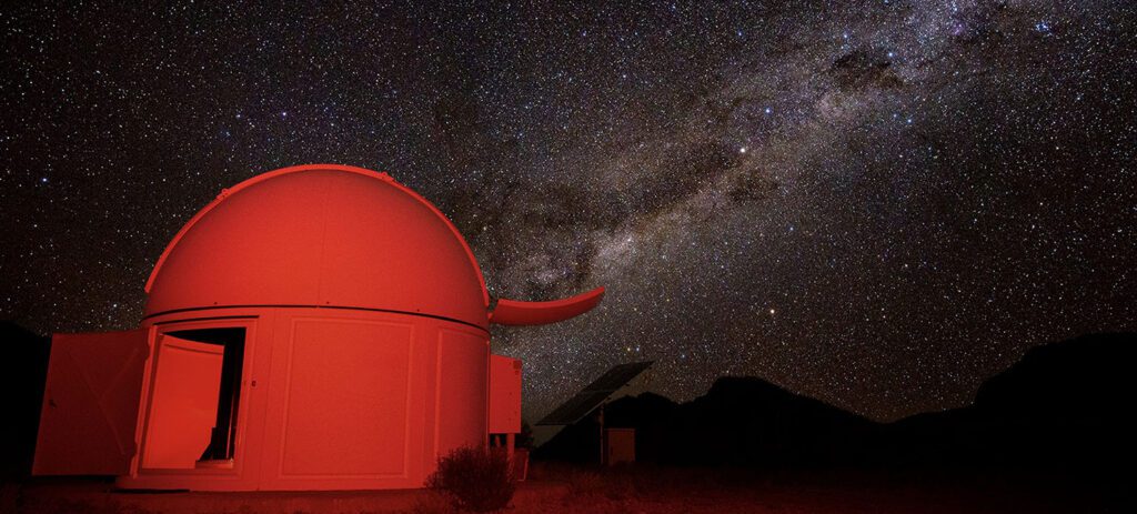 A red-lit observatory dome sits under a dark, star-filled night sky stretching brightly across the horizon above distant silhouettes of mountains. 