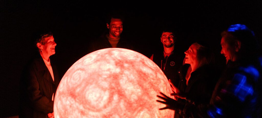 A group of five people stand in a dark room smiling and gathered around a large glowing red sphere that resembles a planet or star, their faces illuminated by its bright light