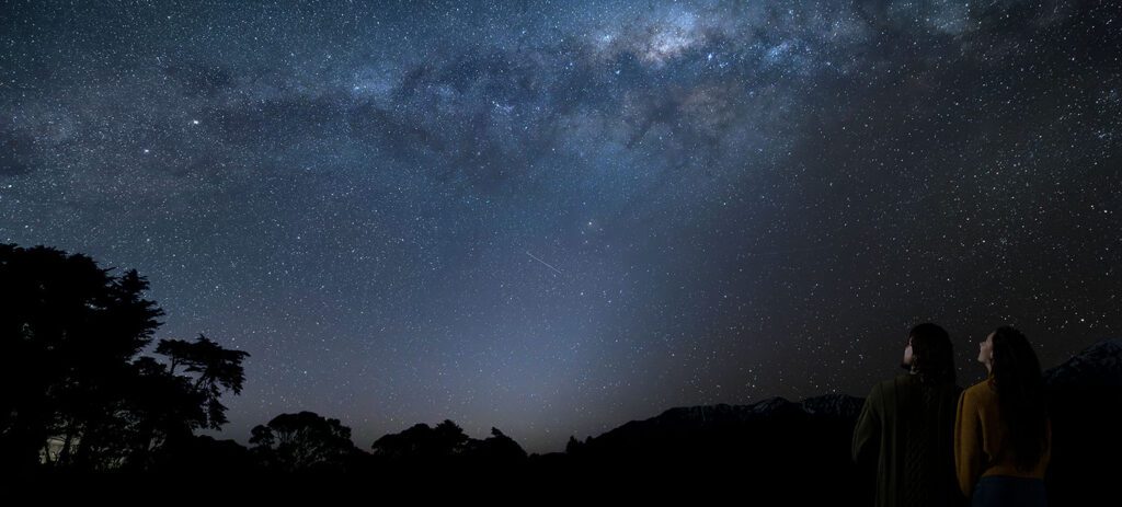 Two people stand together in silhouette at the edge of a dark landscape, looking up at a clear night sky filled with stars and a bright, sweeping band of the Milky Way stretching overhead.