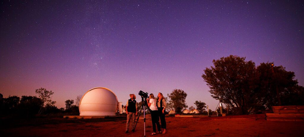 Three people stand together in the red desert looking through a telescope up at a purple-hued night sky filled with stars
