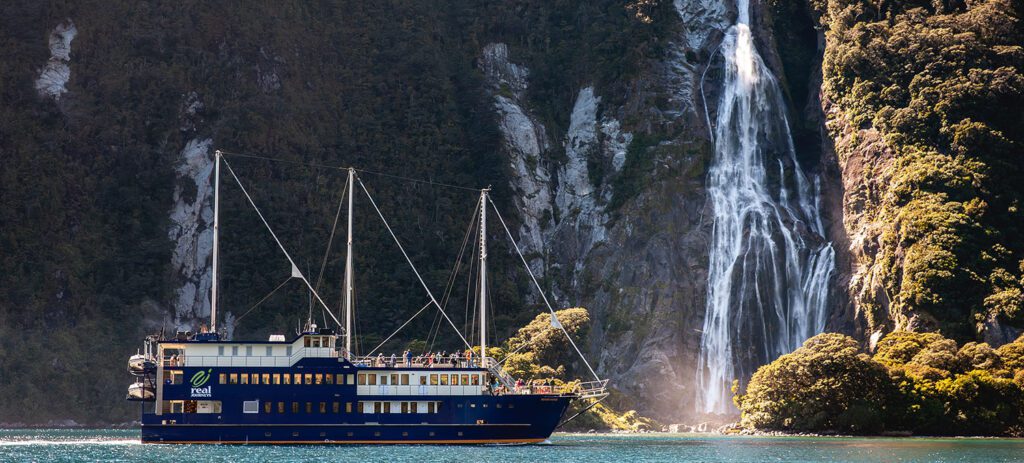 A blue-and-cream cruise ship glides across turquoise water with a tall waterfall cascading down a lush, steep cliff in the background.