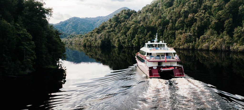 A cruise boat travels through a calm, forest-lined river gorge