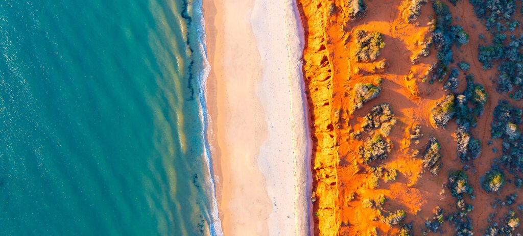 A dramatic aerial shot showing red cliffs meeting white sand and turquoise water. 