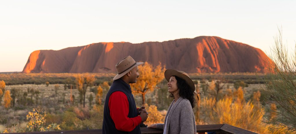 Two people stand facing Uluru at sunset as the rock glows in warm light.