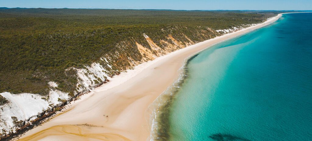 Aerial view of K’gari’s long white sand beach bordered by bright turquoise ocean.