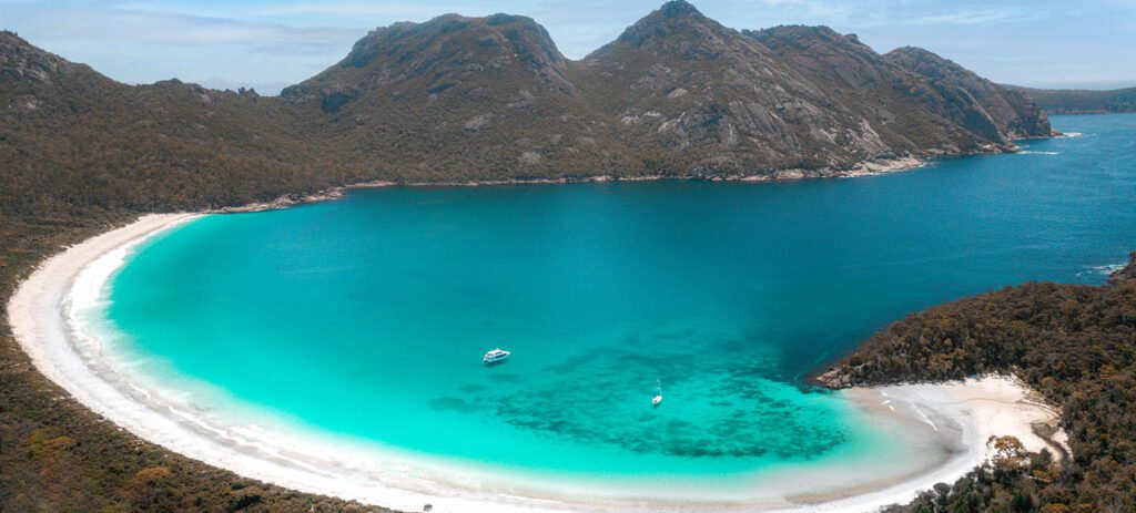 Aerial view of Wineglass Bay featuring a perfect curve of white sand framing bright turquoise water, bordered by forested mountains.
