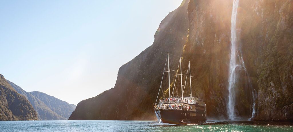 A classic sailing vessel gliding across still water beneath a tall waterfall in Milford Sound, surrounded by steep, sunlit cliffs. 