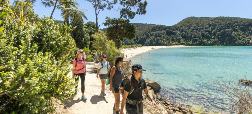 Visitors walking along a coastal track beside turquoise waters in Abel Tasman National Park, Nelson region, New Zealand.
