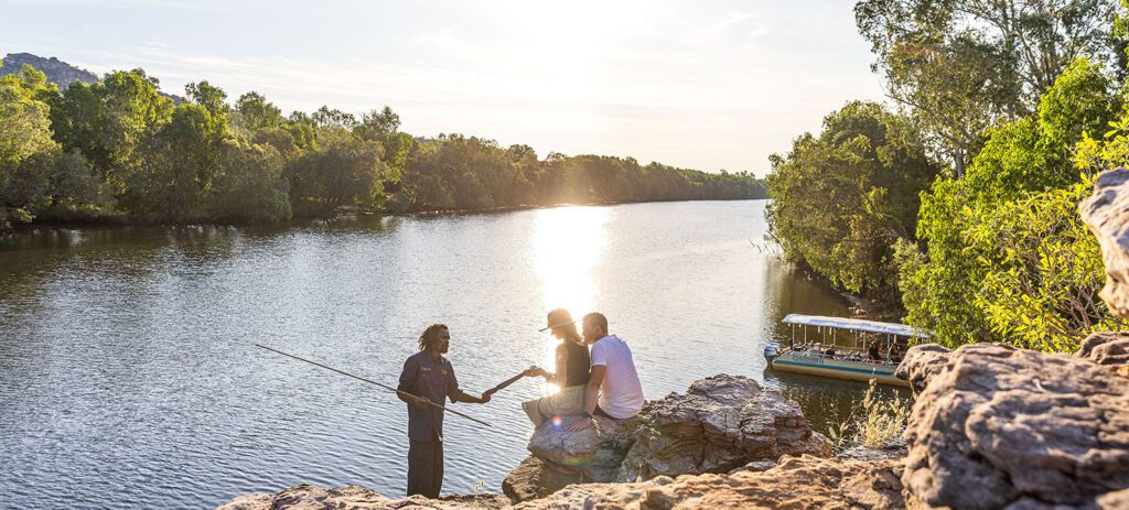 Guluyambi Cultural Cruise guide explaining traditional tools to two guests beside the river, with a cruise boat in the background