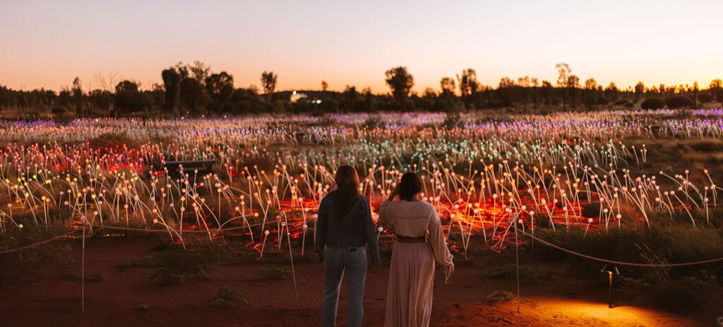 Two people standing in front of the Sunrise Field of Light at Uluṟu, Northern Territory, as the installation glows at dawn.