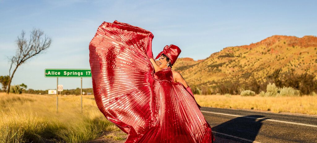 Performer in a flowing metallic red gown posing on a rural highway near a road sign reading “Alice Springs 17,” with desert hills in the background.