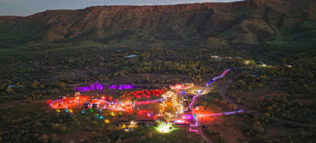 Aerial view of a desert festival at dusk with winding paths outlined in colorful lights and a mountain range rising behind.