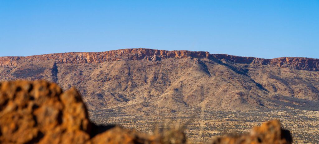 Panoramic view of a rugged desert mountain range with rocky foreground and sparse vegetation under a clear blue sky.