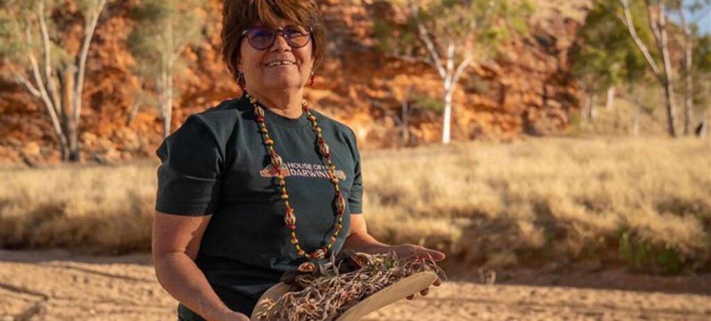 A woman wearing glasses in a black blouse holds an indigenous cooking tool in the Australian Outback