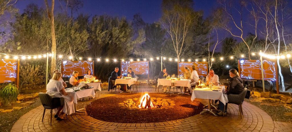 A circular fire pit is in the centre, surrounded by several tables with guests arranged in a semi-circle. Each table is set with white tablecloths, candles, and dinnerware.