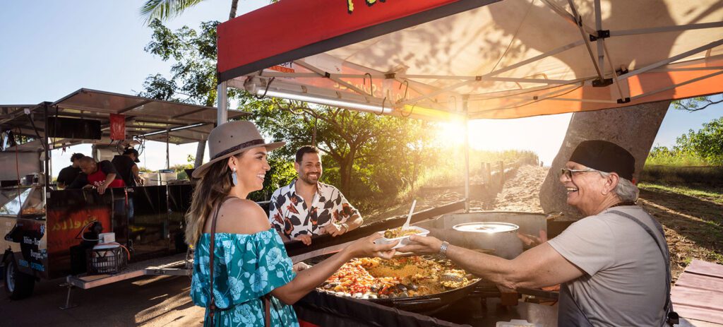 A smiling woman in a blue floral dress and beige hat receives a plate of paella from an older man wearing a black hat and grey shirt at an outdoor food stall. The scene is sunny, with trees in the background, and the stall has a red canopy with the word 'Paella' written on it.