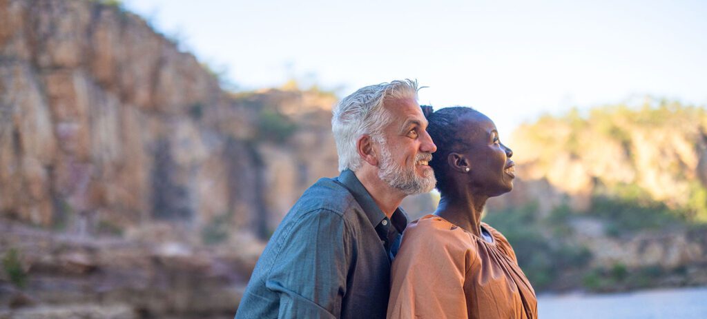 A couple sits closely together by a body of water with rocky cliffs in the background, both gazing upward, the man with grey hair and beard in a blue shirt and the woman in a brown dress with short hair.