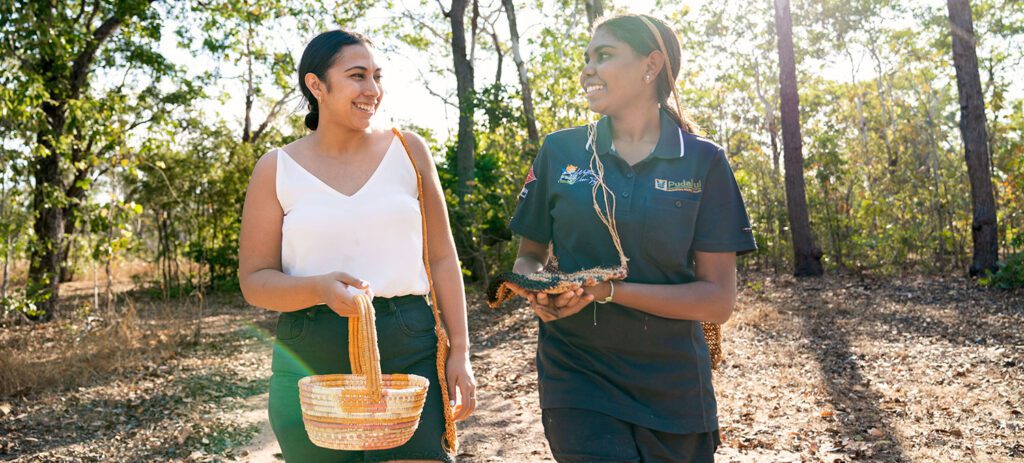 Two women walk through a sunlit forest, smiling and talking while one carries a woven basket and the other holds a handful of natural materials, with tall trees and dappled sunlight in the background.