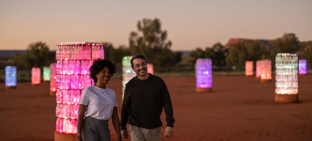 A smiling couple walks across a red desert landscape at dusk, surrounded by glowing cylindrical light installations in shades of pink and purple, with sparse trees and a soft evening sky in the background.