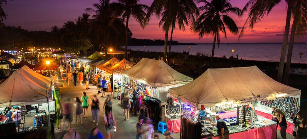 An evening market with colourful stalls and white tents lines a palm-fringed waterfront at sunset, as crowds of people browse under a purple and orange sky.