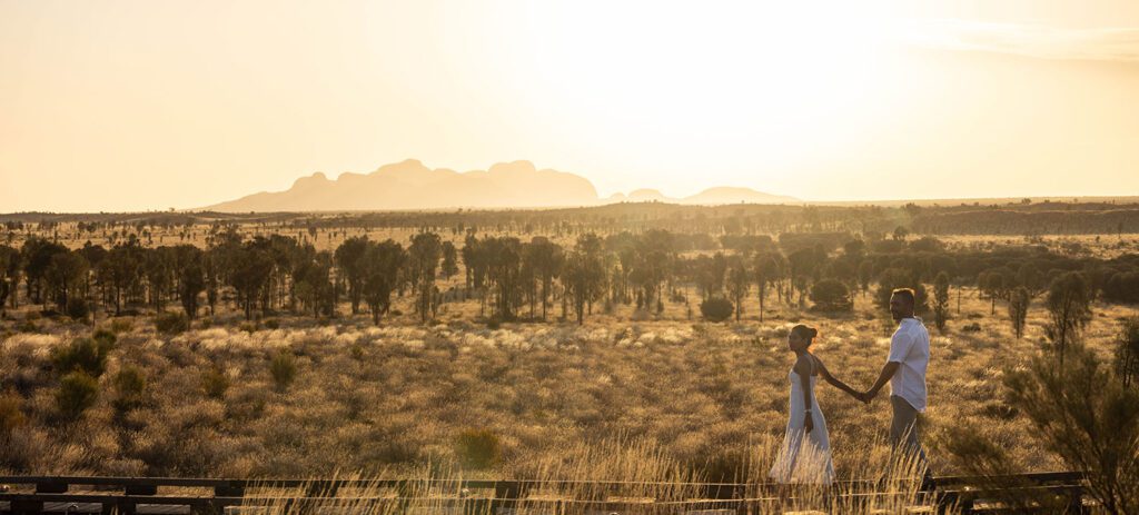 A couple holds hands and walks along a wooden path through golden grassland at sunset, with scattered trees and a distant rocky formation silhouetted against the bright sunsetting sky.