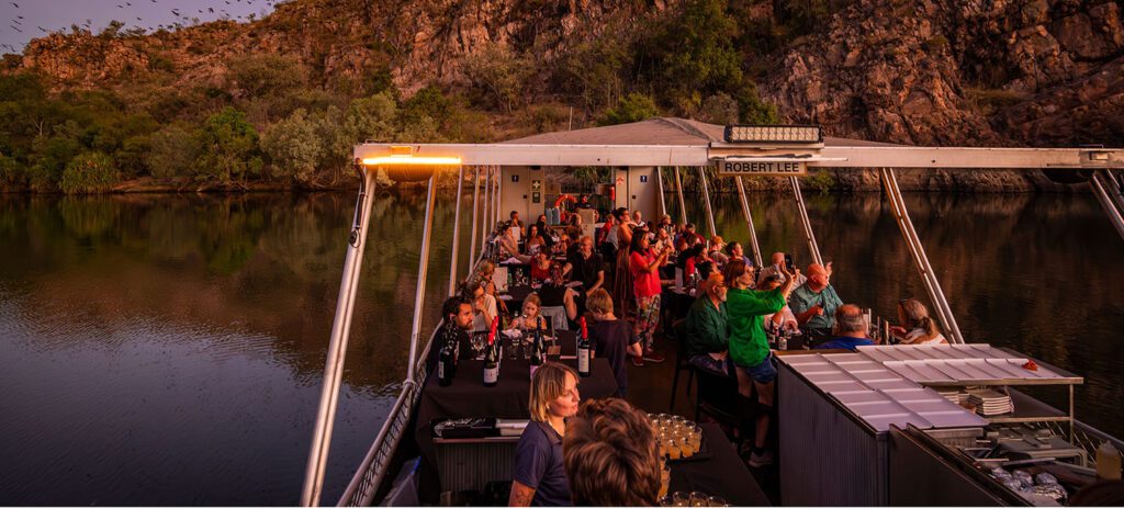 A group of people gather on a boat at sunset along a calm river beneath rocky cliffs, with birds flying overhead