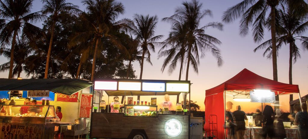 Illuminated food trucks and market stalls under palm trees at dusk with people gathered around outdoor vendors