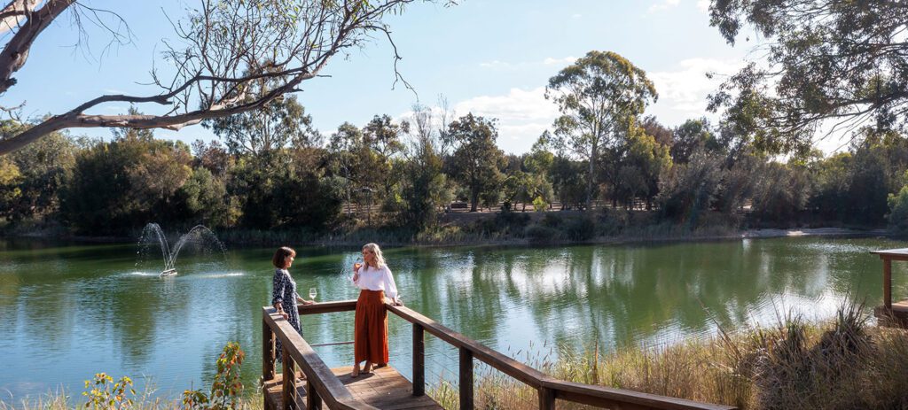 Two people standing on a wooden boardwalk beside a calm lake surrounded by trees and tall grass on a sunny day