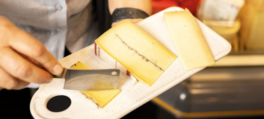 Close-up of a person slicing semi-hard cheese on a white serving board with several wedges displayed for tasting.