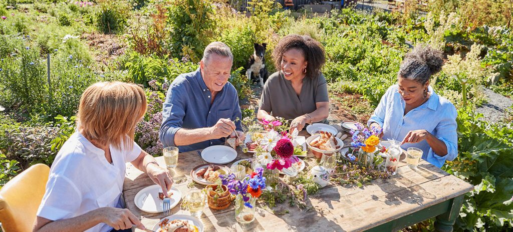 Four adults sharing a meal at a rustic outdoor table surrounded by a lush garden with flowers and farm buildings in the background.