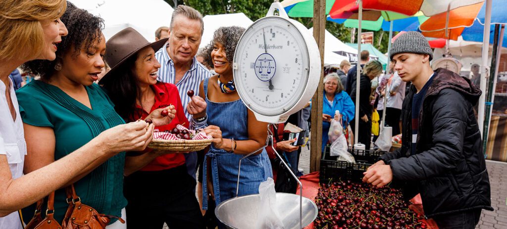 Shoppers sampling fresh cherries at an outdoor market stall with a hanging scale, colourful umbrellas, and bags of fruit on display.