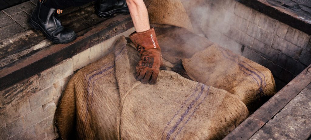 Close-up of a person wearing heat-resistant gloves lifting steaming burlap-covered bundles from a recessed outdoor cooking pit.