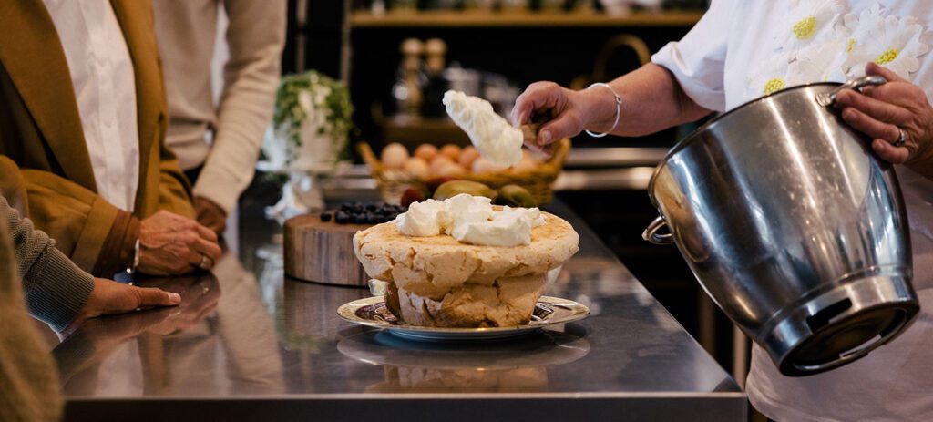 Person adding whipped cream to a layered dessert on a kitchen counter while others watch during a cooking demonstration.