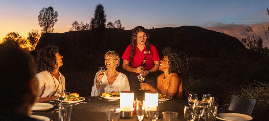 Guests gather around a table, sharing food and wine. A Travel Director pours glasses as the guests dine under the stars beside Uluru