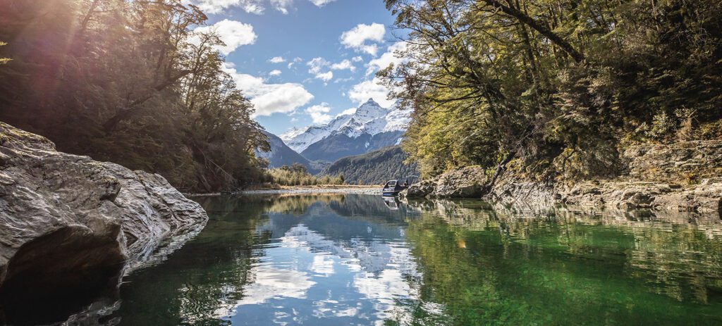 Clear green river reflecting trees and snow-capped mountains under a partly cloudy sky with sunlight filtering through the forest.