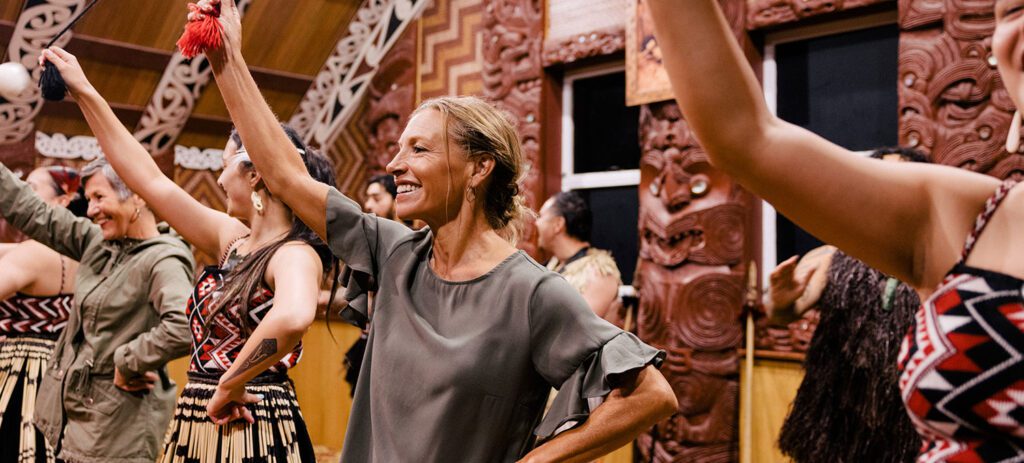 Group of people participating in a Māori cultural performance indoors, smiling and raising poi balls in a carved wooden meeting house.