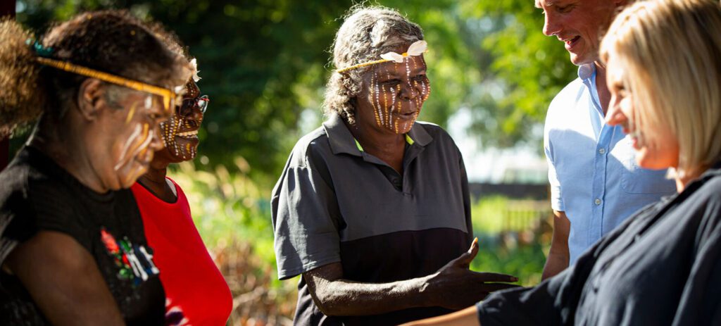 Indigenous hosts with traditional face paint welcome two visitors during an outdoor cultural experience, greeting each other by touching hands in a natural setting.