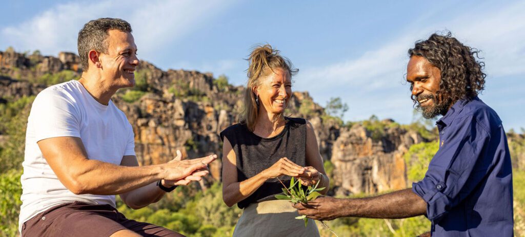 Three people smiling outdoors during a cultural experience as an Indigenous guide holds leafy branches, with rugged sandstone cliffs and bushland in the background.