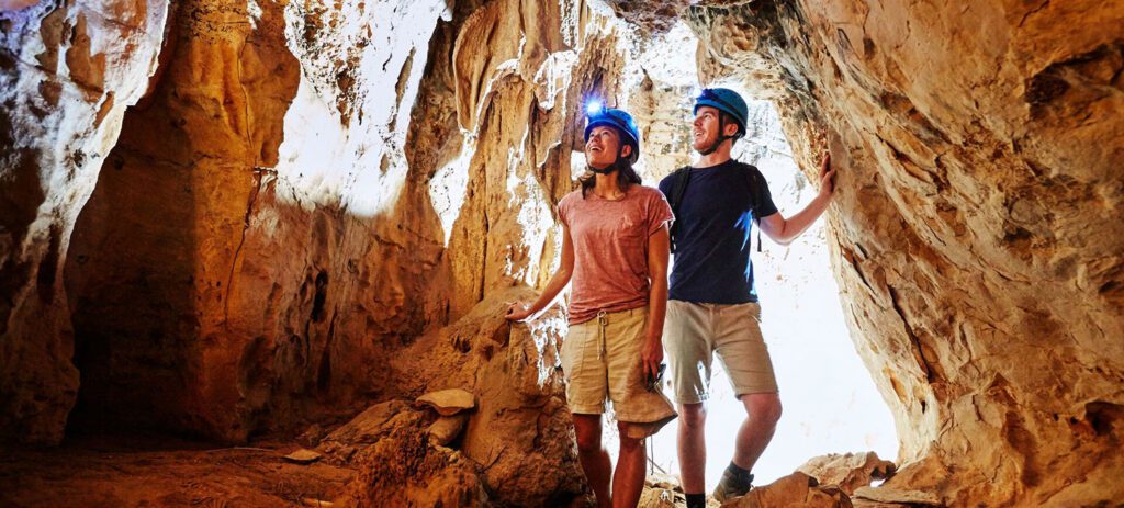 Two people wearing helmets with headlamps explore a limestone cave, looking up at illuminated rock formations and stalactites.