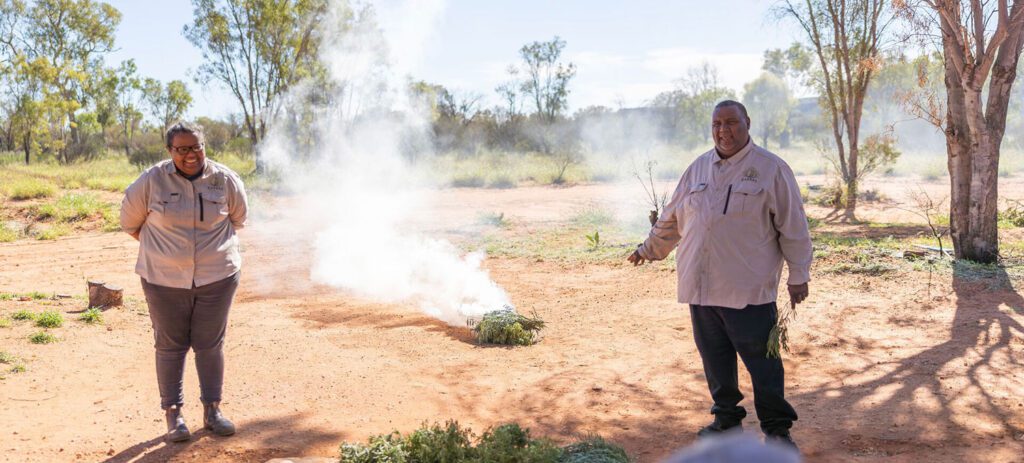 Two Indigenous guides lead an outdoor cultural demonstration around a smoking fire with native plants, while visitors watch in a bushland setting.