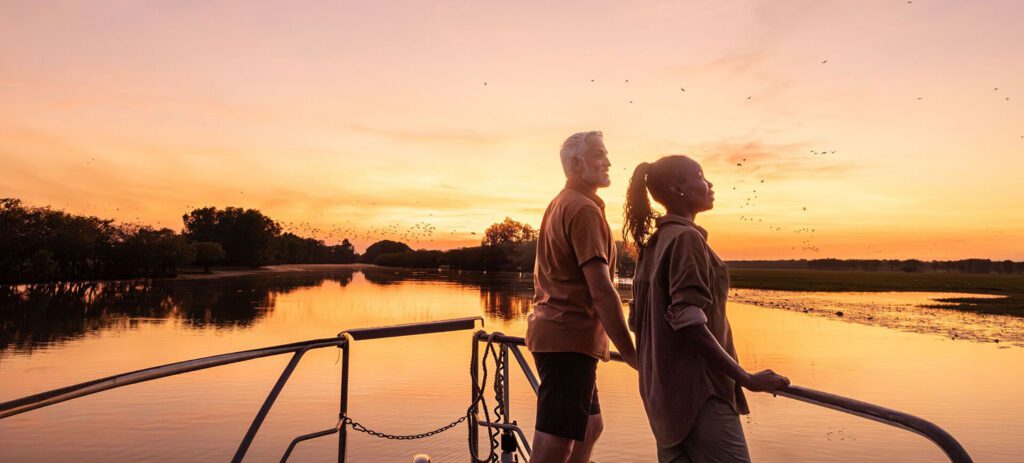 Two people stand at the bow of a boat watching birds over a calm wetland at sunset, with golden reflections on the water and a colourful evening sky.