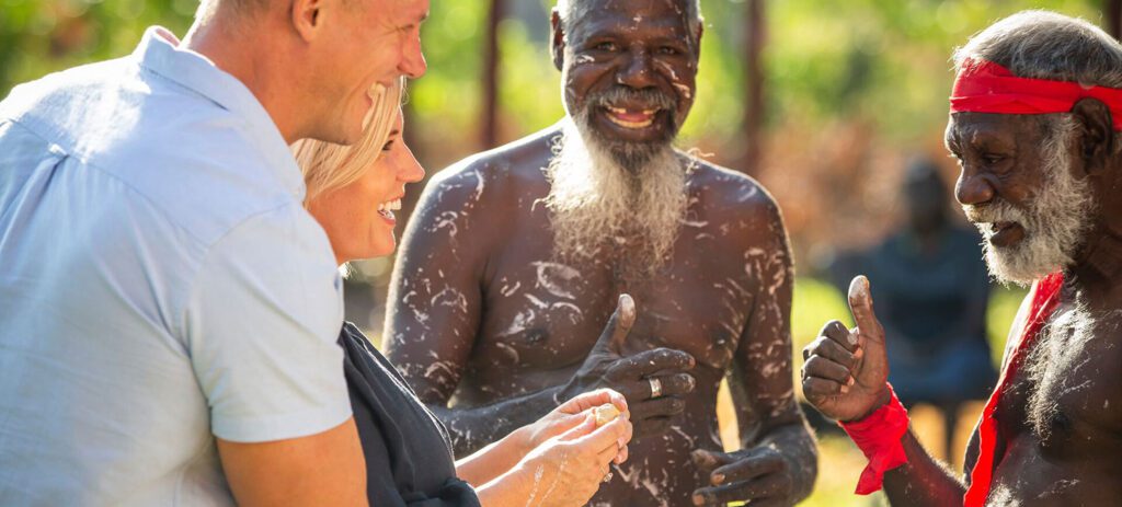 Two visitors smile while sharing food with Indigenous elders during an outdoor cultural experience, with traditional body paint, red ceremonial armbands, and a woven basket visible.