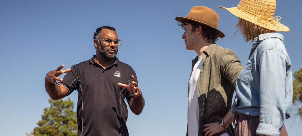 Indigenous guide gestures while explaining cultural knowledge to two visitors outdoors, with the group listening in a sunny bushland setting.