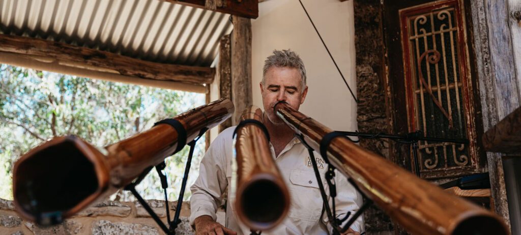 Musician performs on multiple didgeridoos in a rustic indoor venue, with the traditional instruments mounted on stands in the foreground.